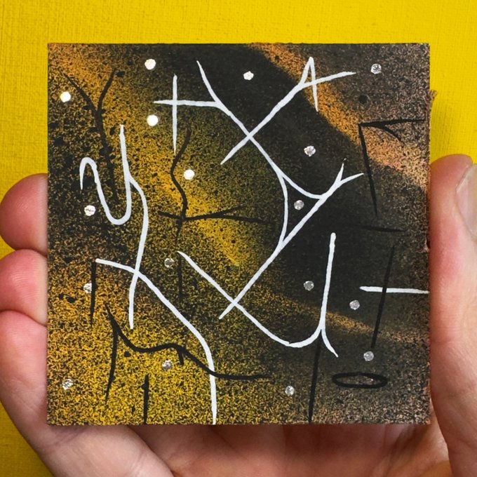 The image shows a hand holding up a small square art piece, which appears to be a painting of stars and lines. The artwork has a black background with white lines and dots, giving it an abstract and artistic appearance. The person's fingers are visible on either side of the frame, emphasizing the size and importance of the artwork being displayed.