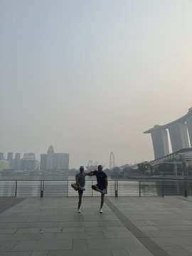 Photo of two people standing on a waterfront walkway taking a selfie, with the Marina Bay Sands hotel and Singapore skyline visible in the hazy background.
