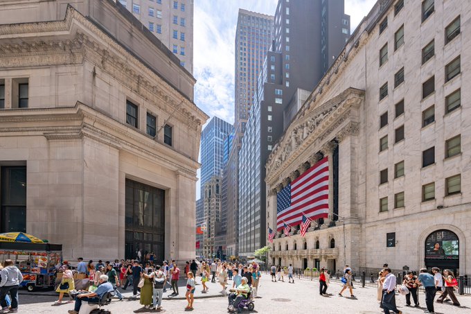 A street-level photograph of the New York Stock Exchange building on Wall Street, featuring its iconic large American flag hanging between the columns, with pedestrians walking on the sidewalk and tall skyscrapers in the background on a sunny day.