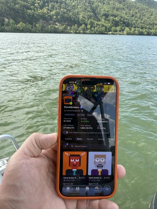 A hand holding a smartphone showing an NFT marketplace (StonkBrokers) with prices in ETH, displayed against a lake and forested mountains backdrop.