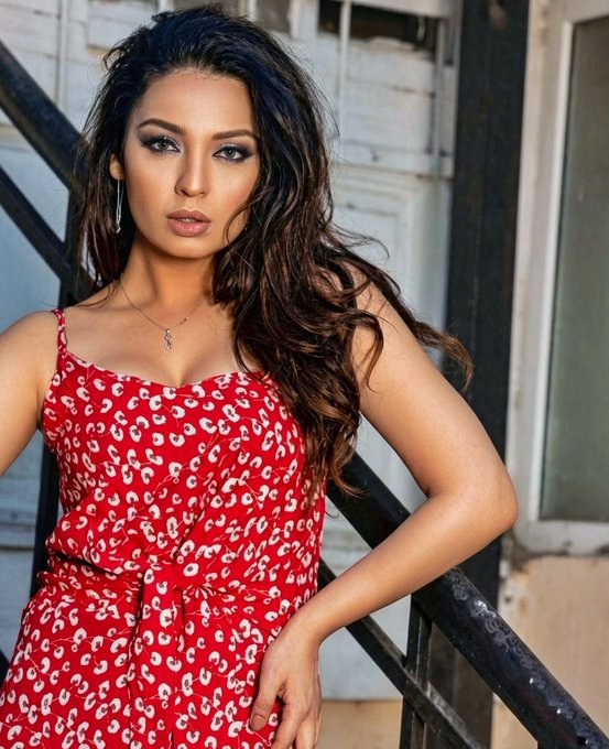 A woman with long dark wavy hair poses in a red patterned sundress with white floral designs, leaning against a metal railing in an industrial-style setting.