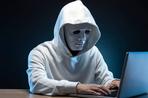 A person wearing a white hoodie and a featureless white mask sits at a desk typing on a laptop in a dark setting with blue lighting.