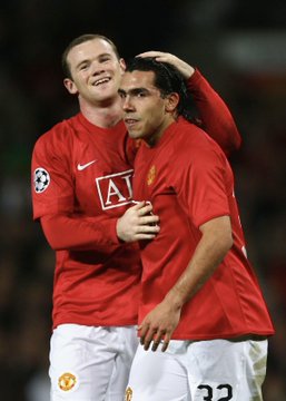 Two Manchester United footballers embrace in celebration while wearing red jerseys with the "A1" sponsor logo and the number 33 visible on his shorts, with Rooney commenting on Tevez's passionate but limited-English playing style during their Champions League travels together.