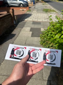 A hand holds three promotional cards for 'MYRIAD STOCK', each featuring a QR code and an illustration of a person, with a paved sidewalk, greenery, and parked cars in the background.