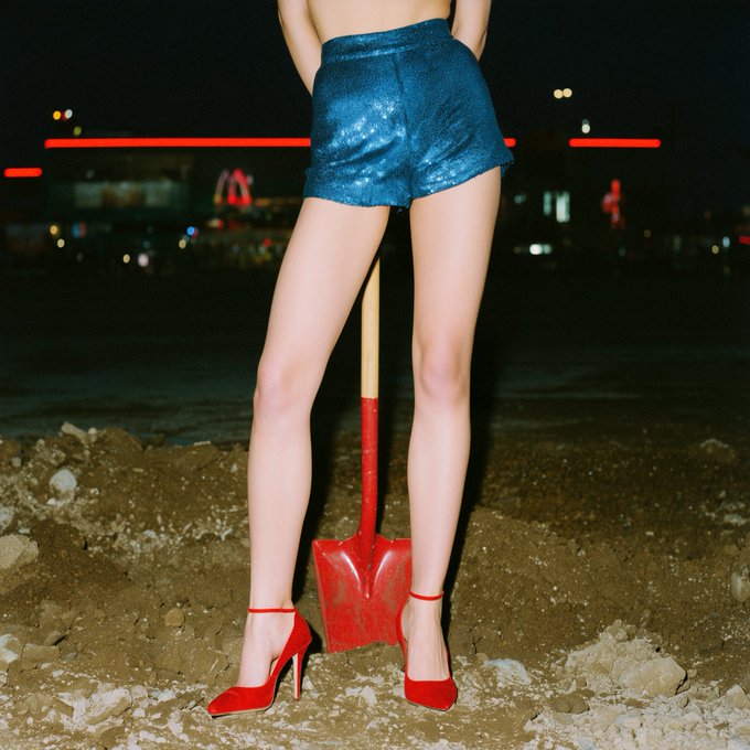 A person stands on dirt wearing sparkly blue shorts and red heels, holding a red shovel between their legs against a nighttime city backdrop.