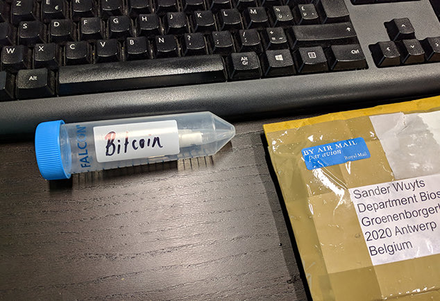 A Falcon tube labeled "Bitcoin" sits beside a keyboard and an airmail envelope addressed to Sander Wuyts at the Department of Bioscience in 2020 Antwerp, Belgium—likely depicting one of the physical prizes that geneticist Nick Goldman distributed after encoding a Bitcoin into DNA in January 2015.