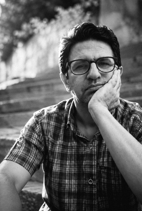 A black-and-white portrait of a man wearing glasses and a plaid shirt, resting his chin on his hand with a contemplative expression.
