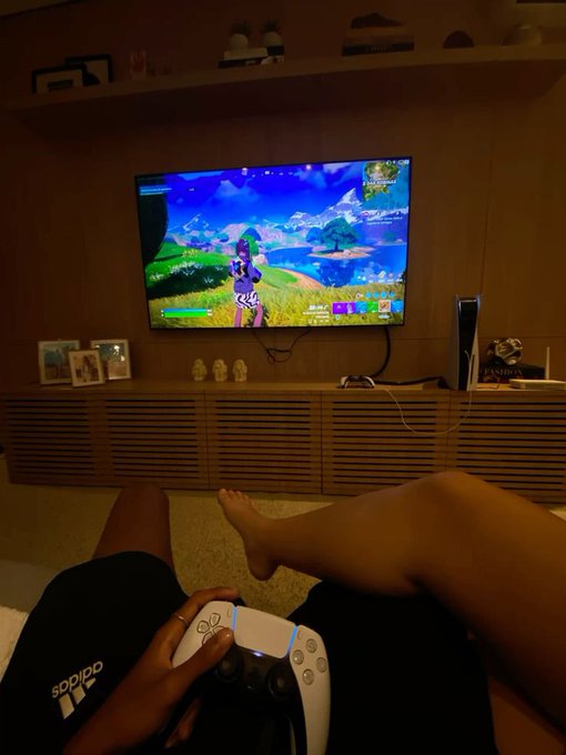 A person relaxing on the floor playing Fortnite on a PlayStation 5, with the game displayed on a wall-mounted TV and the player's bare feet and Adidas shorts visible in the foreground.