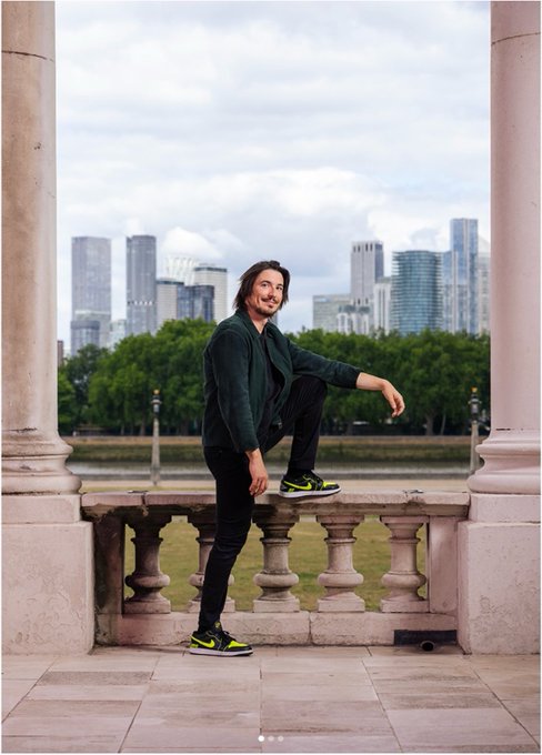 A professional photograph of a young man with long hair wearing a dark green blazer, black pants, and neon yellow sneakers, posing with one foot on a stone balustrade with a modern city skyline in the background.