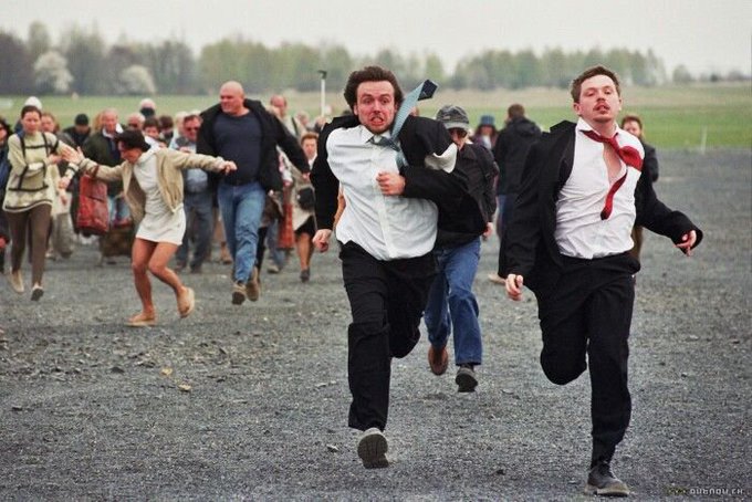 A humorous staged photograph showing a crowd of people in business attire running frantically across a gravel lot, with two men in white shirts and dark suits in the foreground appearing panicked.
