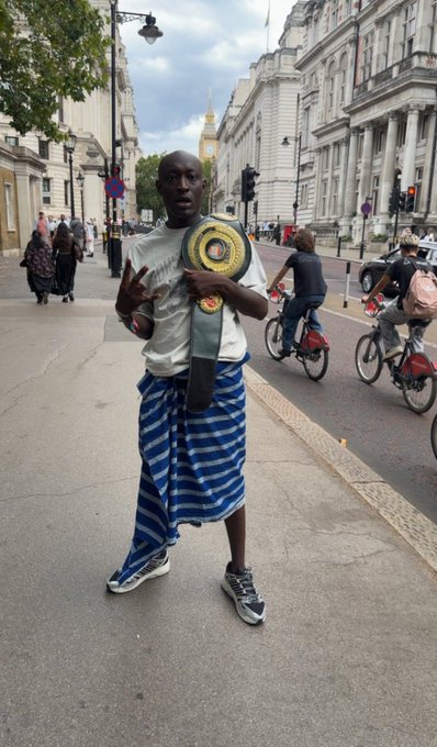 A man wearing a striped sarong, white shirt, and gold championship belt standing on a London street with classical buildings and cyclists in the background.