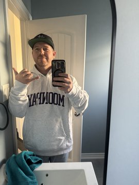 A man taking a mirror selfie wearing a grey Moose Knuckles hoodie and a black baseball cap, holding a phone.