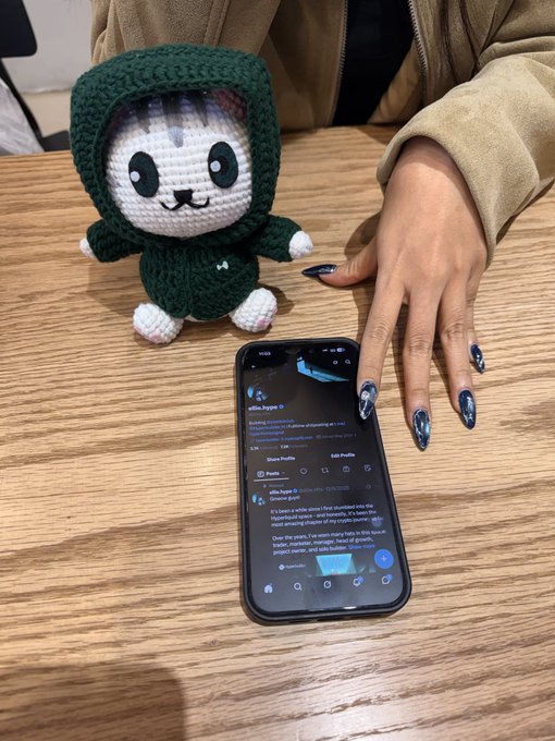 A hand with long, decorated nails rests on a wooden table next to a smartphone displaying a Twitter profile and a small crocheted plush toy wearing a green hooded outfit.