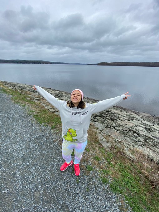 A woman in a gray hoodie and colorful leggings standing with arms outstretched in a celebratory pose by a lake under cloudy skies.