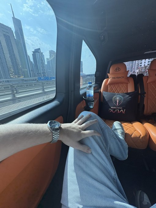A person wearing a watch and light blue jeans relaxes inside a luxury SUV, with a "MAYBACH" branded pillow reading "SPECTACULAR DUBAI" visible across the cabin, while a skyline of skyscrapers is seen through the window. The "GM GM GM" post suggests a luxurious morning in Dubai, capturing the vibe of a stylish "good morning" from inside a high-end ride.