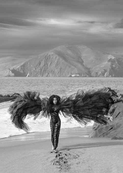 A surreal black-and-white photograph of a woman in a patterned bodysuit standing on a beach, wearing dramatic, expansive dark wings that blend into the seascape, with mountains rising across the water behind her.