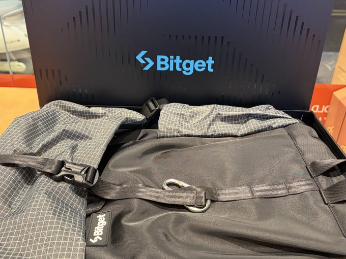 An open Bitget-branded box containing a black drawstring bag with a Bitget label and a checkered gray-patterned item, possibly a backpack or bag.