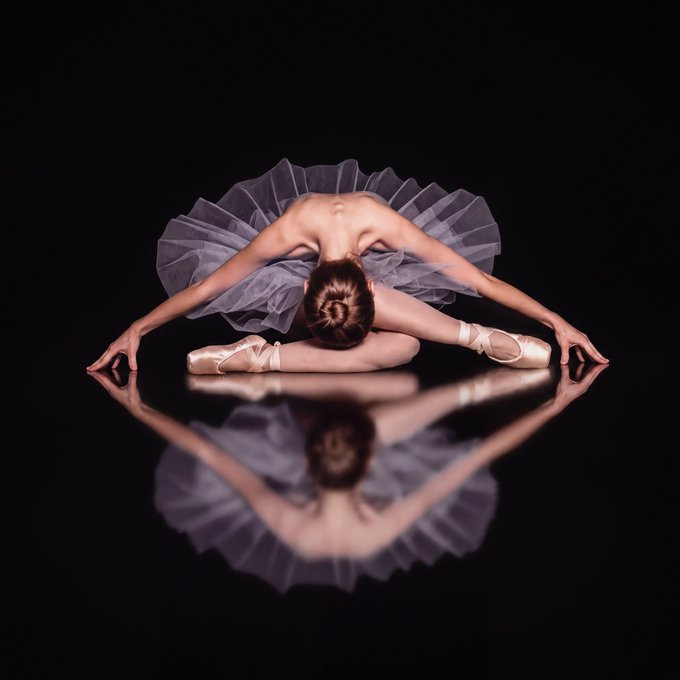 A ballerina in a gray tutu and pointe shoes performs a forward fold pose on a reflective surface, creating a mirror image against a black background.