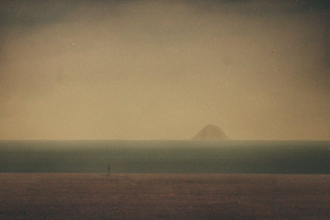 A vintage-style, grainy landscape photograph showing a lone figure standing in a vast open field with a distant mountain or hill under a muted, sepia-toned sky.