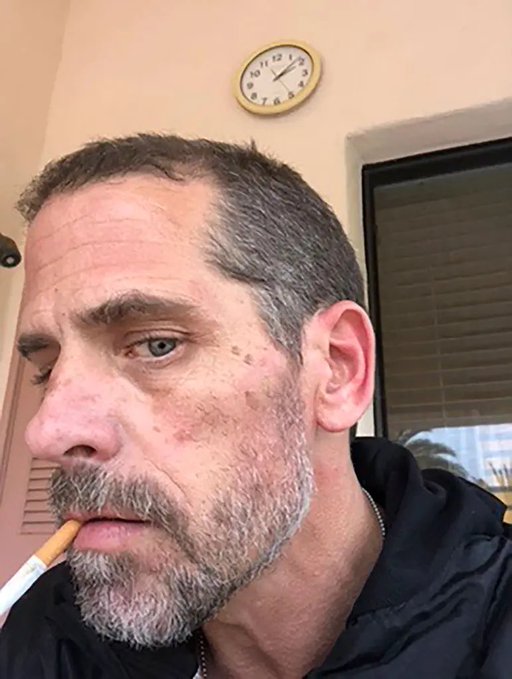 A close-up selfie shows a gray-bearded, weathered man with a cigarette hanging from his lips, staring blankly off-camera beneath a wall clock reading roughly 12:18. The gritty image is paired with a post mocking people as "Junkies and Crackheads" launching memecoins and defending Hunter Biden, suggesting the photo is meant to visually embody the "crackhead" insult used in the caption.