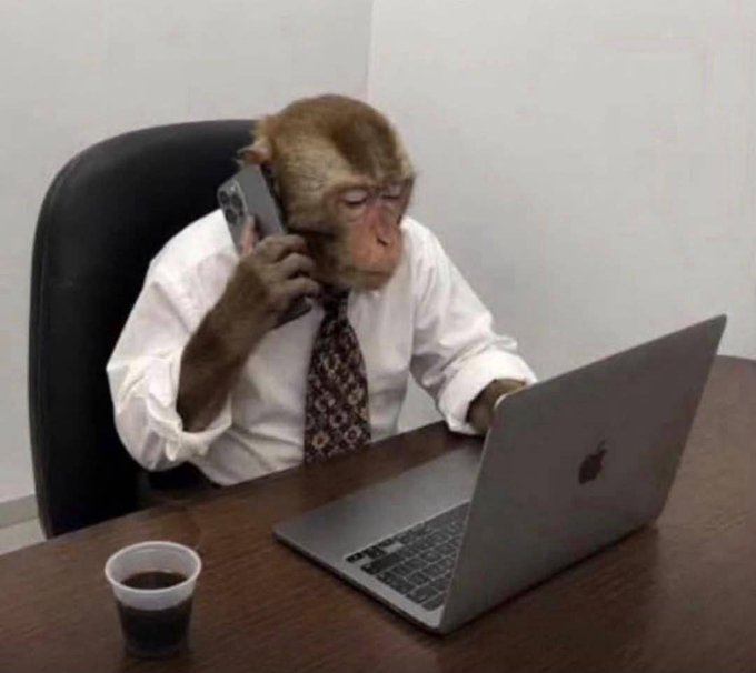 A humorous meme image of a monkey dressed in a white business shirt and tie, sitting at a desk while talking on a mobile phone and working on a silver laptop (appearing to be an Apple MacBook), with a coffee cup beside it.