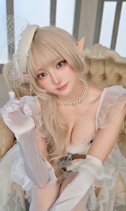 A young woman with long blonde hair, pointed elf-like ears, and green eyes poses elegantly in a white frilly outfit with long satin gloves and a pearl choker, evoking a fairy-tale aesthetic. The image aligns with the "Chira" theme, suggesting a stylized cosplay or character portrayal of a delicate, ethereal figure.