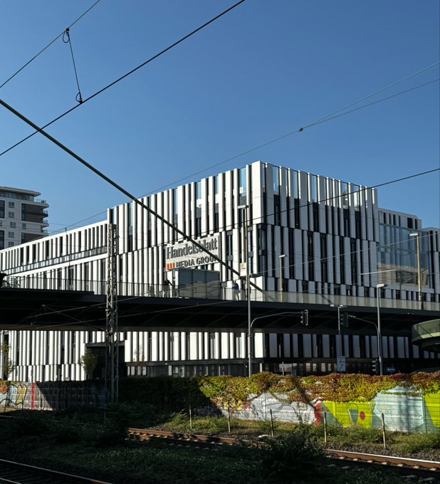 A modern, multi-story office building with vertical slatted facade panels bearing 'Handelsblatt' and 'DIE MEDIA GRO' signage, photographed under a clear blue sky with overhead tram wires crossing the frame.