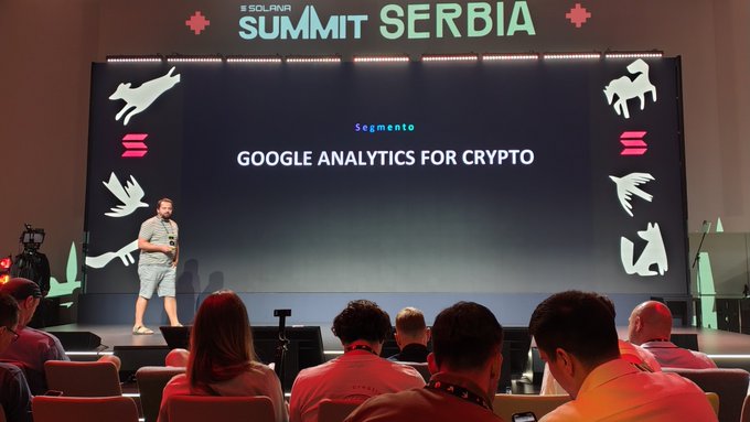 A presenter stands on stage at 'SOLANA SUMMIT SERBIA,' giving a talk titled 'GOOGLE ANALYTICS FOR CRYPTO' to an attentive audience.