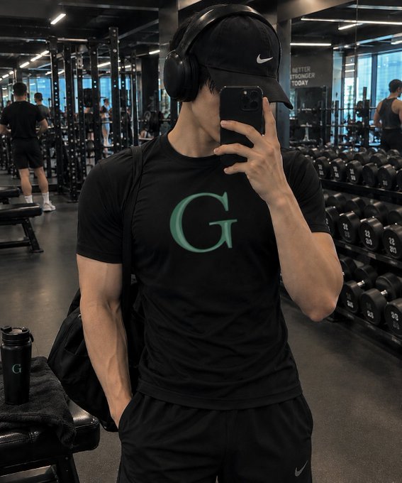 A muscular man takes a gym selfie wearing a black Nike cap, headphones, and a black t-shirt with a teal 'G' logo, with dumbbells visible in the background.