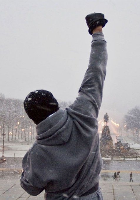 A person in a gray hoodie and beanie stands triumphantly with an arm raised high, hat held aloft in a snowy landscape featuring a statue in the background. The image captures a moment of personal victory and determination, visually reinforcing the post's message about how discipline—not just talent—is what leads to winning.