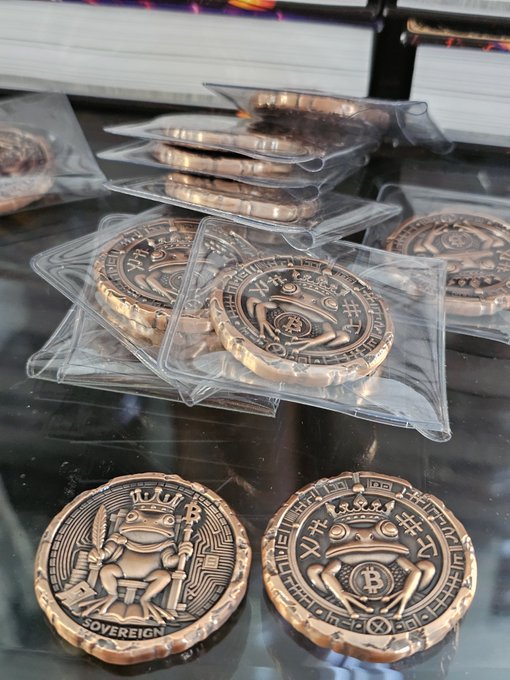A close-up photo displays several copper "SOVEREIGN" coins featuring a crowned frog holding a scepter, stacked in clear plastic sleeves on a dark reflective surface.