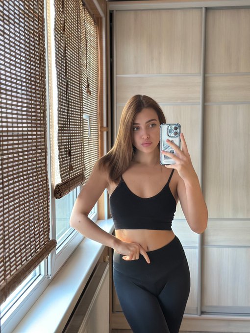 A woman in a black sports bra and leggings takes a mirror selfie with her phone in a room with wooden closets and bamboo blinds.