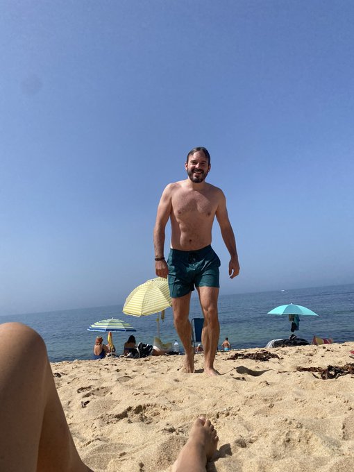 A man with a beard wearing teal swim trunks stands barefoot on a sandy beach, smiling at the camera, with the ocean, striped umbrellas, and other beachgoers in the background.