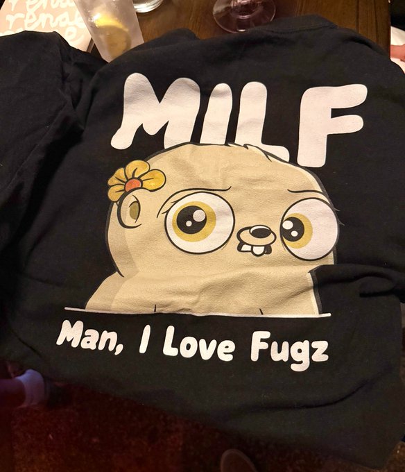 A black t-shirt featuring the Chimpers character (a wide-eyed cartoon monkey with a yellow flower) is displayed on a table, with bold text reading "MILF - Man, I Love Fugz" above and below the illustration, highlighting the collaboration between Chimpers and Fugz mentioned in the post.