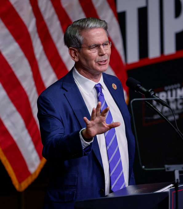 A man with gray hair and glasses, wearing a blue suit and purple striped tie, speaking at a podium with an American flag in the background and partial text 'TRUMP' and 'TRUM' visible.