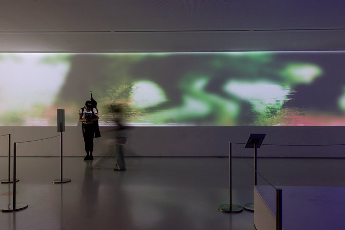 A woman stands alone in a dimly lit gallery space, silhouetted against a massive illuminated media wall displaying blurred, dreamlike green and red imagery. The artwork, titled "To Be Perceived, Against Evil" by Linda Dounia and Rheaplex, is shown as part of the Schlosser Media Wall at Moving Image NYC, complementing the post's invitation to Ana María Caballero's "Material and Materiality" presentation.