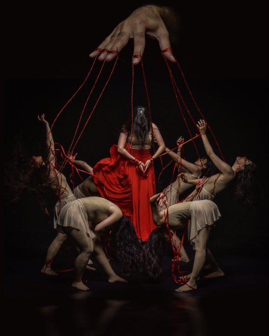 A dramatic photograph depicts dancers in flowing dresses connected by red strings to a giant hand reaching down from above, set against a black background.