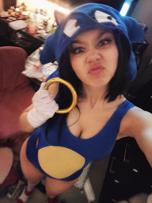 A woman poses for a selfie wearing a blue Sonic the Hedgehog cosplay outfit, complete with a hood featuring ears and a gold ring prop.