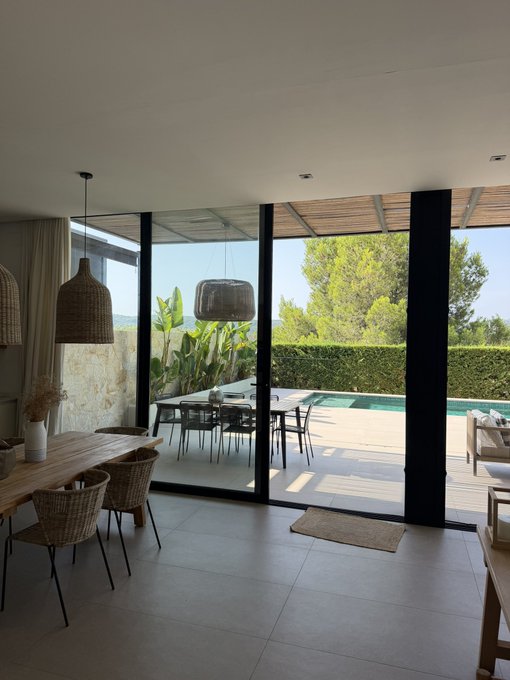 An interior view of a modern home with a dining area featuring a wooden table and wicker chairs, looking out through large glass doors to a patio with a swimming pool and lush greenery.