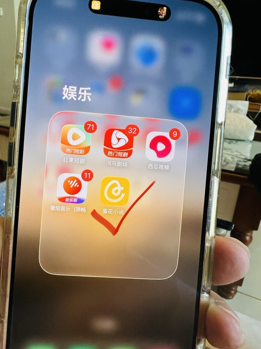 A hand holds up an iPhone displaying a folder labeled "娱乐" (Entertainment) with Chinese apps including 热/剧 (71 notifications), 热门剧榜 (32 notifications), 西瓜视频 (9 notifications), 番茄音乐 (11 notifications), and 蜜花小说, with a hand-drawn red checkmark circling the 蜜花小说 app, suggesting the author is highlighting a lesser-known app that defies the mainstream trading/FOMO trend discussed in the post.