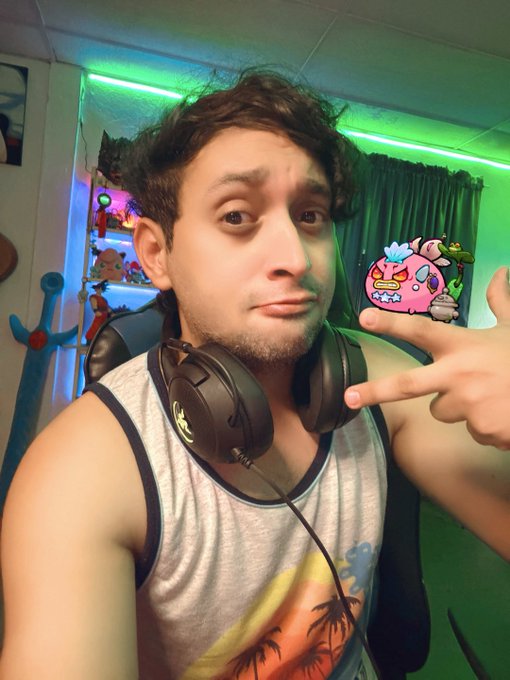 A selfie of a young man wearing a gray tank top and Razer headphones, making a peace sign, with green ambient lighting and a small Angry Birds character graphic in the background.