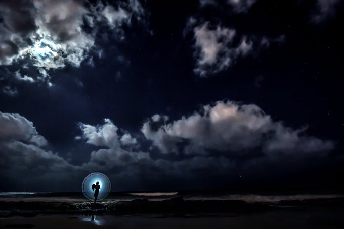 A nighttime photograph of a person standing on a rocky shoreline inside a glowing circular portal or light effect, under a dramatic cloudy sky lit by the moon.