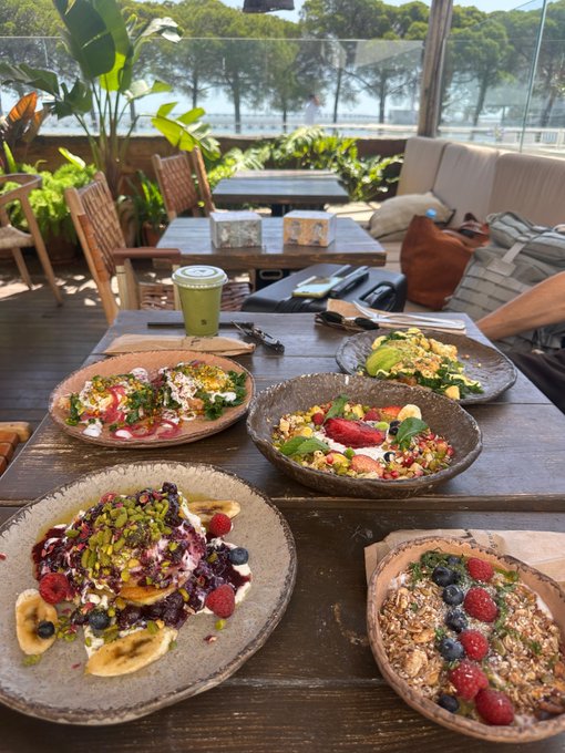 A photograph of a wooden table on an outdoor restaurant patio featuring four colorful, healthy dishes including salads and smoothie bowls topped with fruits, nuts, and seeds, with a green juice and avocado toast in the background.