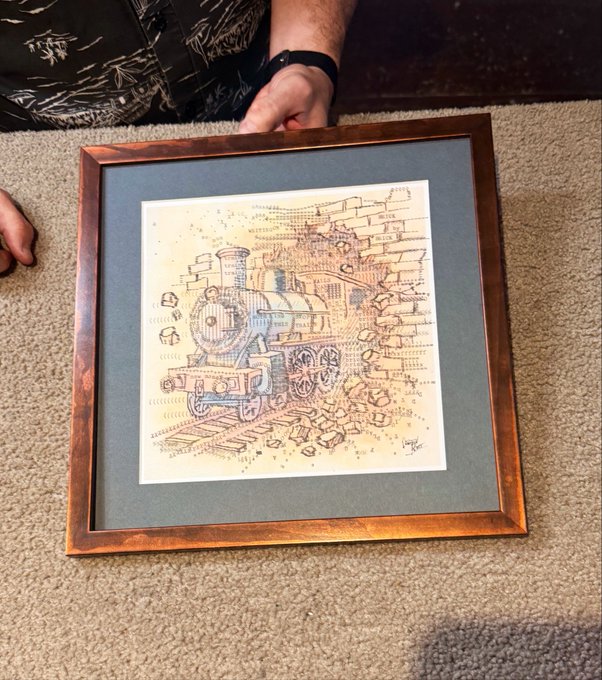 A framed hand-drawn illustration of a steam locomotive on aged parchment paper, resting on a carpeted floor with a person's hand visible at the top edge.