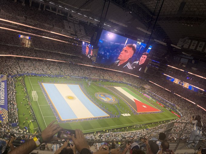 A packed stadium during the FIFA World Cup 2026, with giant flags of Argentina and Jordan spread across the field and players shown on the jumbotron, as fans in the foreground capture the spectacle on their phones—a stark contrast to the post's "Infiltrating the opps" caption, which seems unrelated to the celebratory pre-match atmosphere.