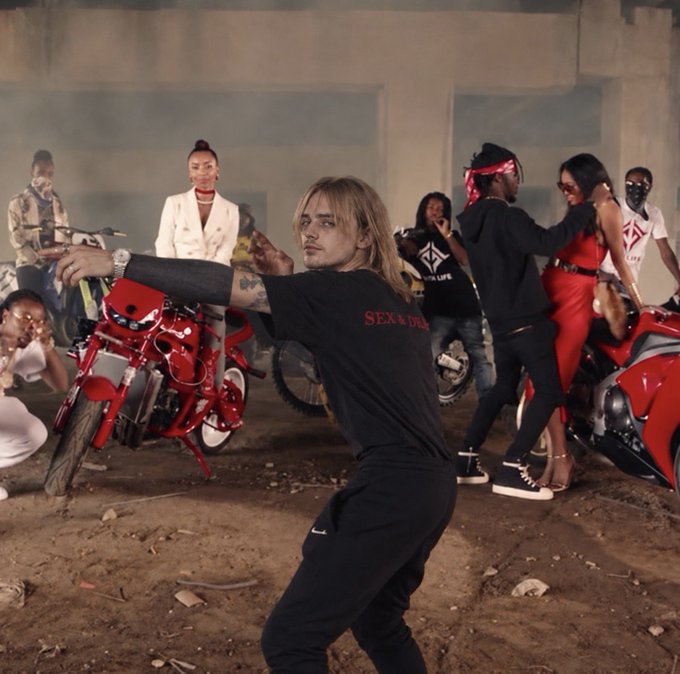 A music video still showing a man with blonde-streaked hair in a black 'SEX' t-shirt dancing in front of a group of people and red motorcycles at night.