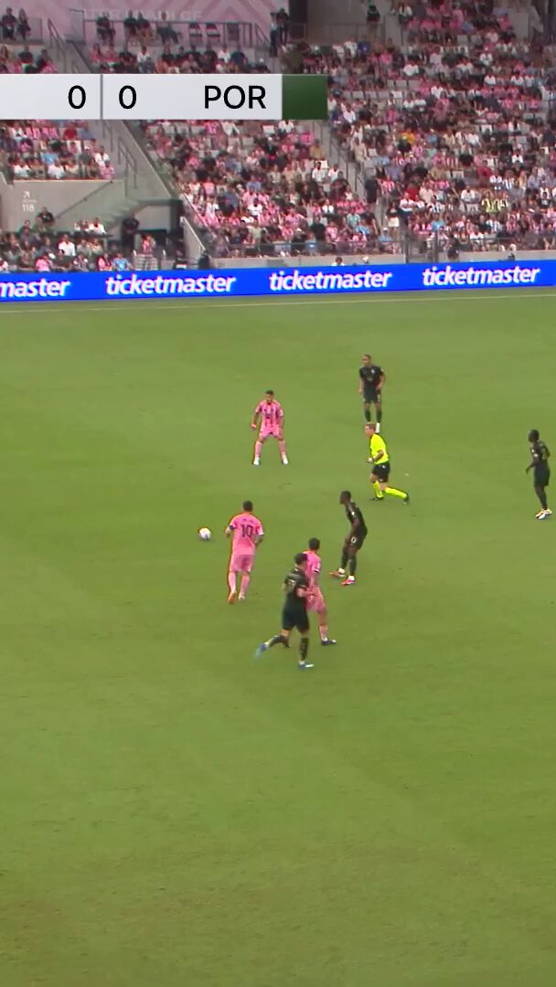 The image shows a group of people playing soccer on a field with a pink advertisement banner behind them. The players are wearing colorful uniforms and actively participating in the game, which appears to be taking place at a stadium or sports event. The photo is taken from an aerial perspective, providing a clear view of the action on the field.