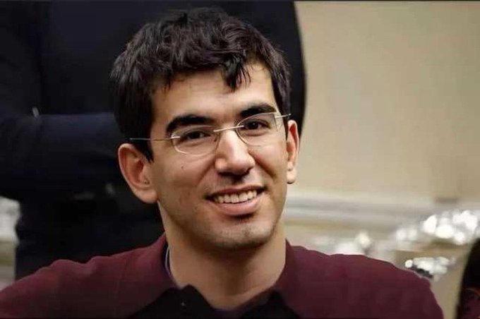 A close-up photograph of a smiling young man with dark hair and glasses wearing a maroon shirt.