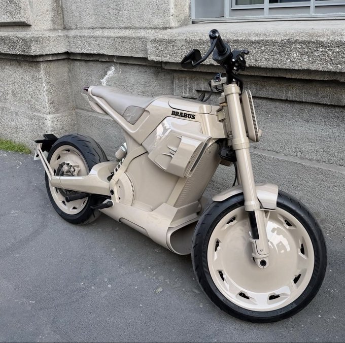 A cream/tan-colored futuristic motorcycle with "BRABUS" branding on the tank is parked on a paved surface in front of a stone building. The bike features distinctive solid disc-style wheels with cutouts and an angular, modern design.