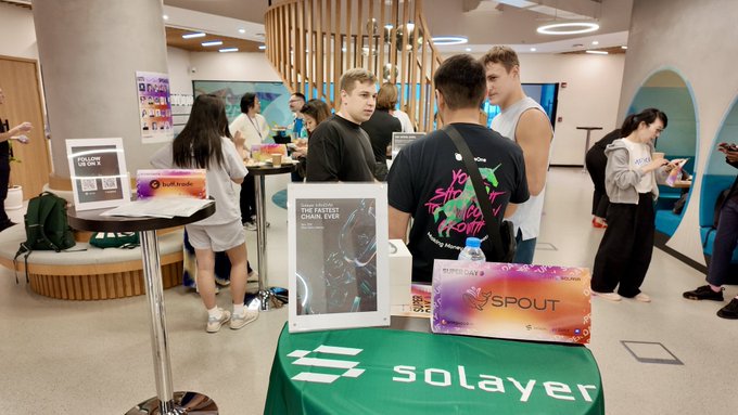 An indoor event/meetup scene showing people gathered around tables with booths for crypto projects including 'solayer' (with green branding) and 'SPOUT', with attendees networking in a modern venue.